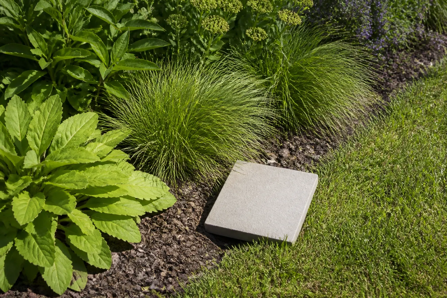 Een lichte steenstaal ligt op de rand van een gazon en een brede border met herhaalde siergrassen en vaste planten.