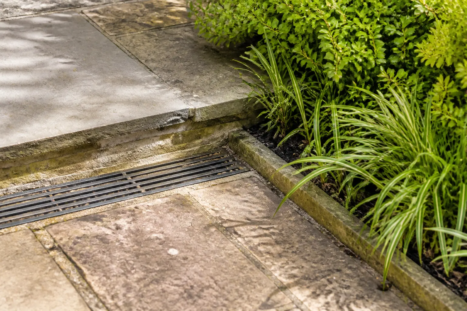 Een smalle afvoergoot en lage trede liggen waar verweerde bestrating aan een beplante tuinrand grenst.