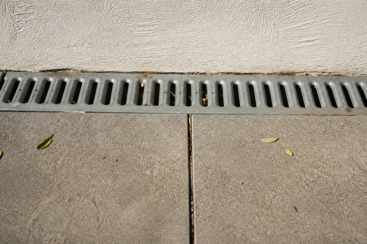 Old paving meets a metal drain and rendered wall in a close view, giving clear lines for checking whether a generated image has shifted the hardscape.