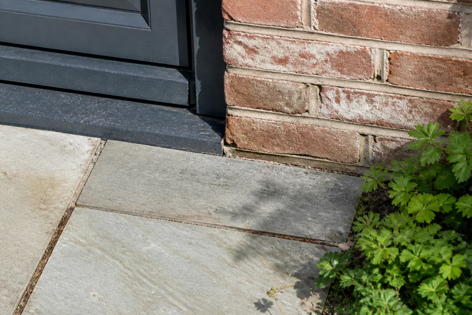 A brick house wall, door threshold and paving edge meet in a tight photograph, showing fixed features an AI garden render should preserve.