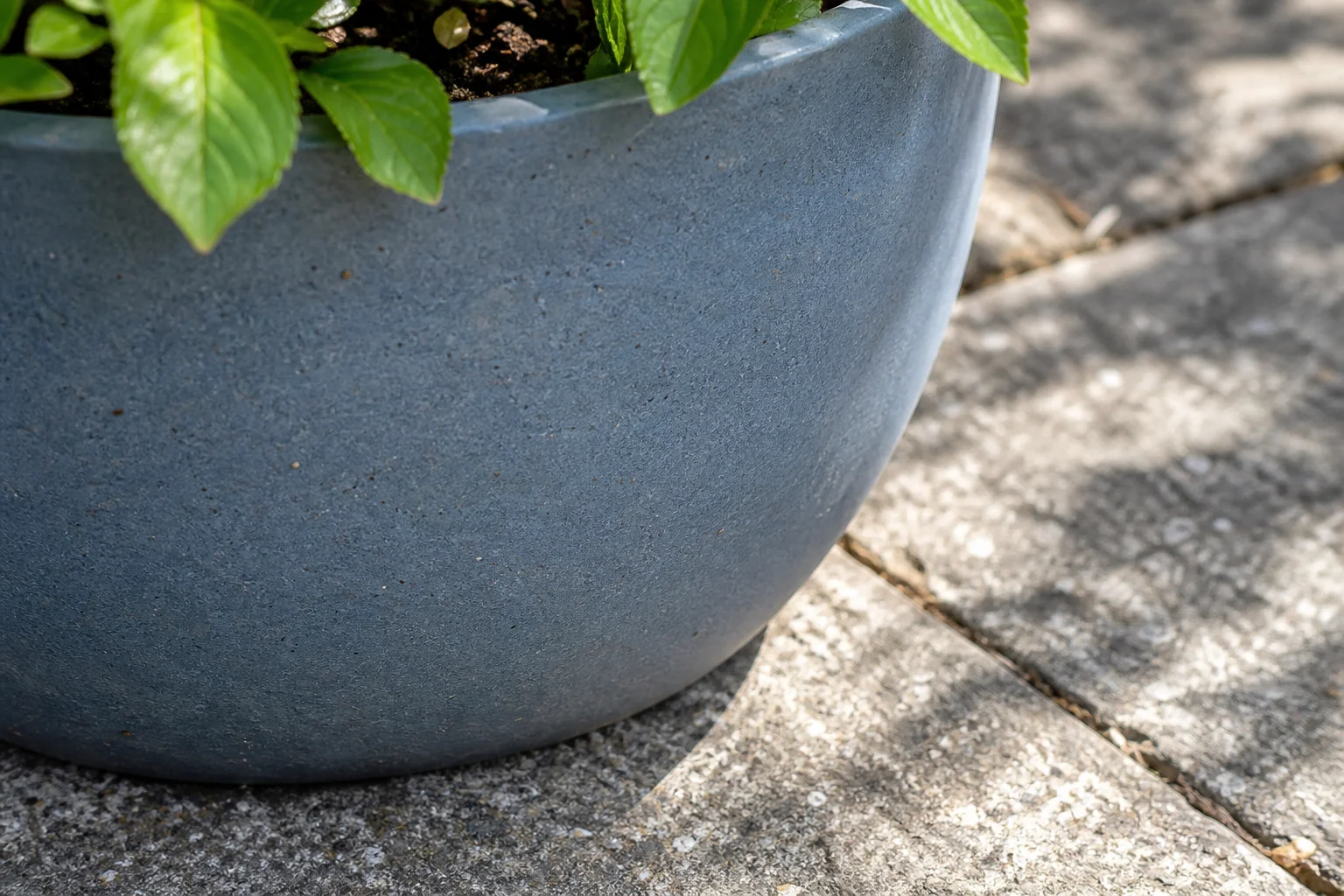 Een matte blauwgrijze plantenpot staat op gespikkelde grijze tegels, met groene bladeren boven de rand.