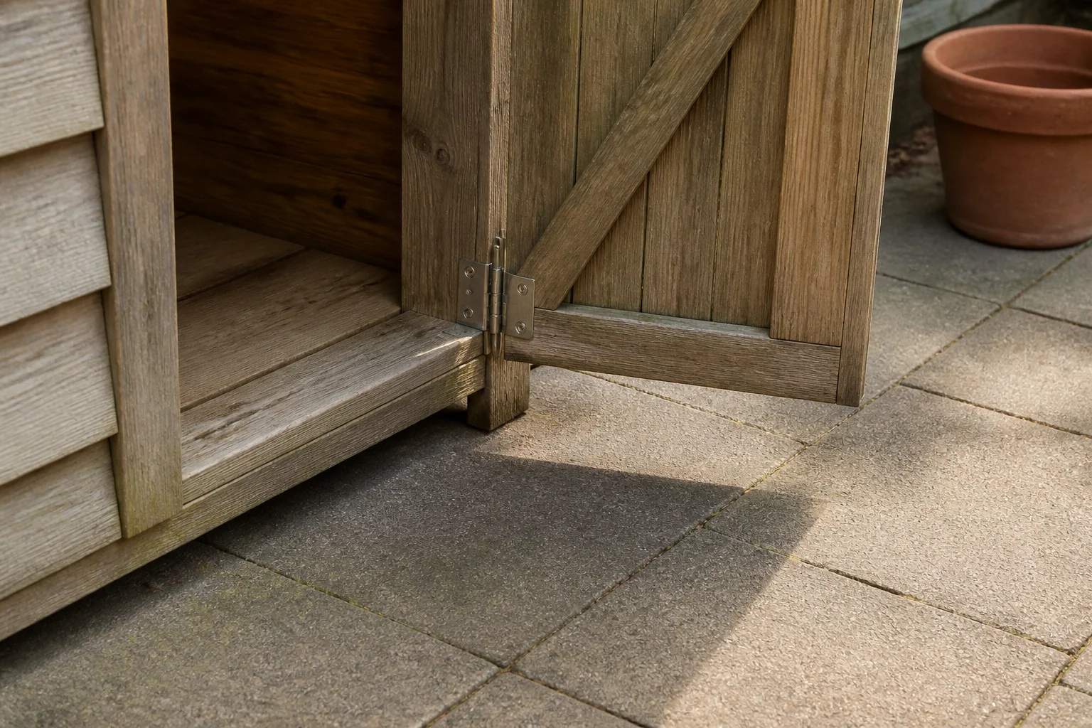 The lower door of a timber garden cupboard stands open above paving with clear space beside it.