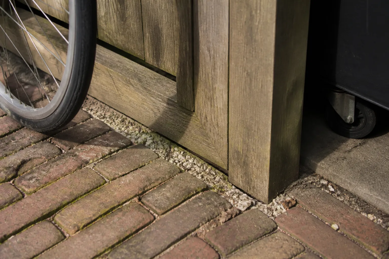 A bicycle wheel and bin caster have clear space beside a weathered timber screen, brick paving and a gravel drain