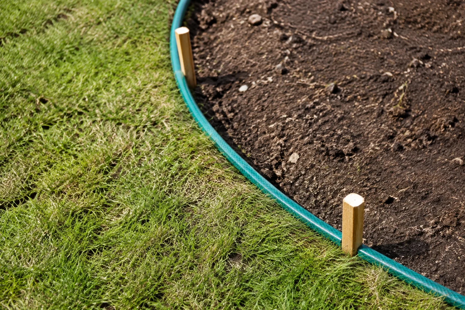 Een groene tuinslang en twee eenvoudige houten paaltjes markeren een deel van een gebogen border tussen gebruikt gazon en kale grond.