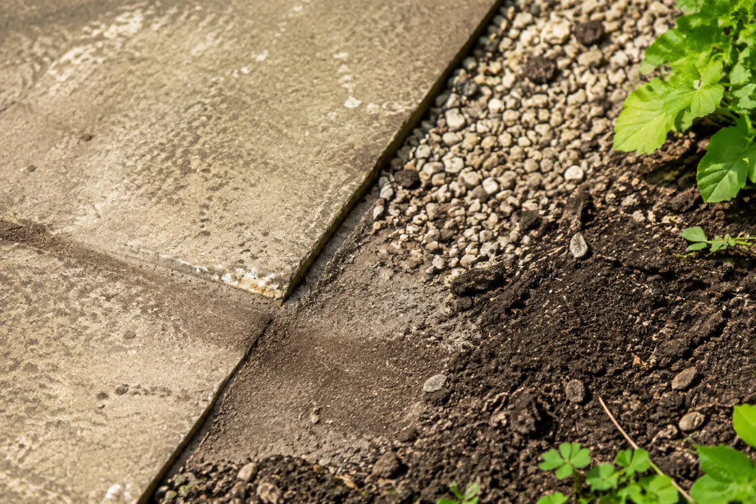 Een verweerde tegel sluit aan op grind en aarde, waar lichte slibsporen een mogelijke route van afstromend water tonen.
