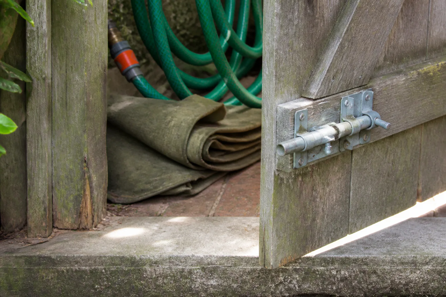 A weathered wooden gate stands open beside a coiled hose and folded protective canvas at a narrow threshold.