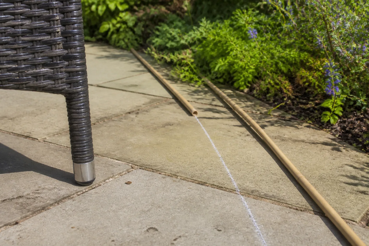 A chair foot, a chalk sightline and two bamboo markers align across worn garden paving towards a planted boundary.