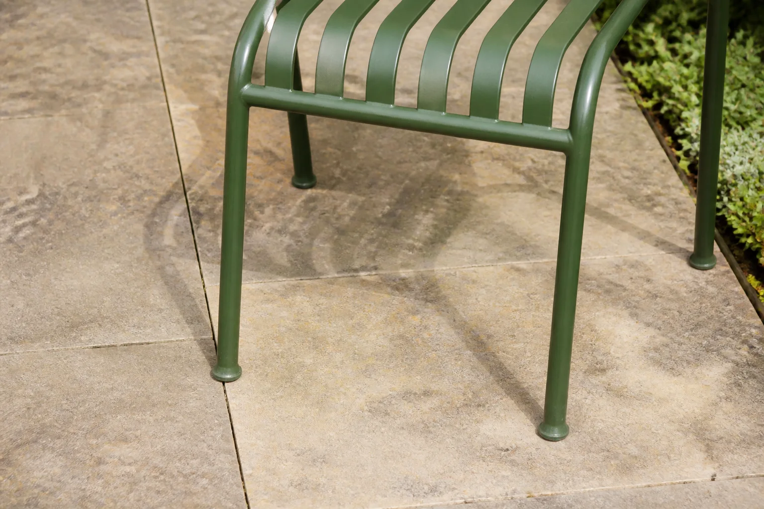 Green metal chair feet stand on worn pale paving with open ground beside them.