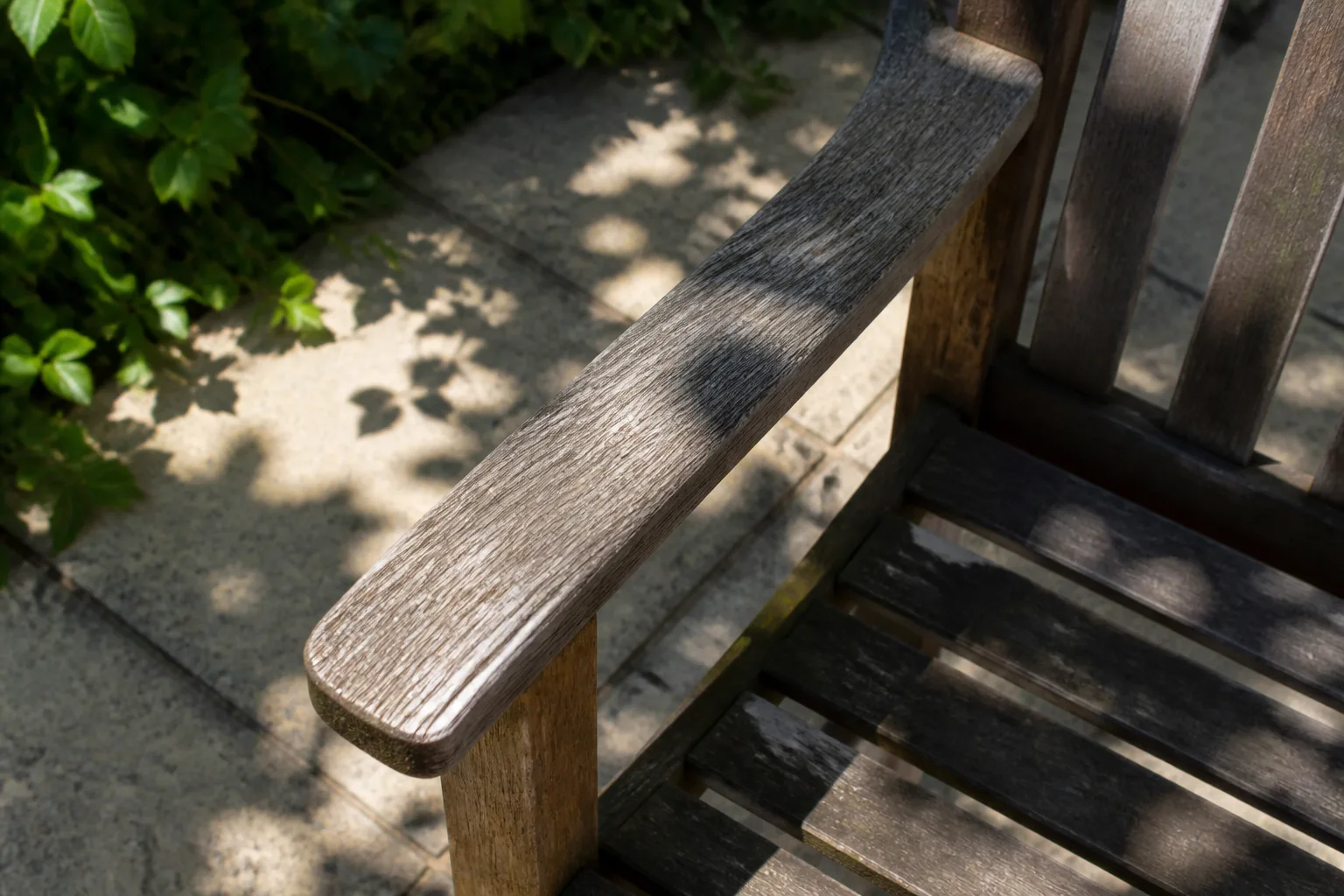 Leaf shadows fall across the weathered arm and slats of an outdoor chair.