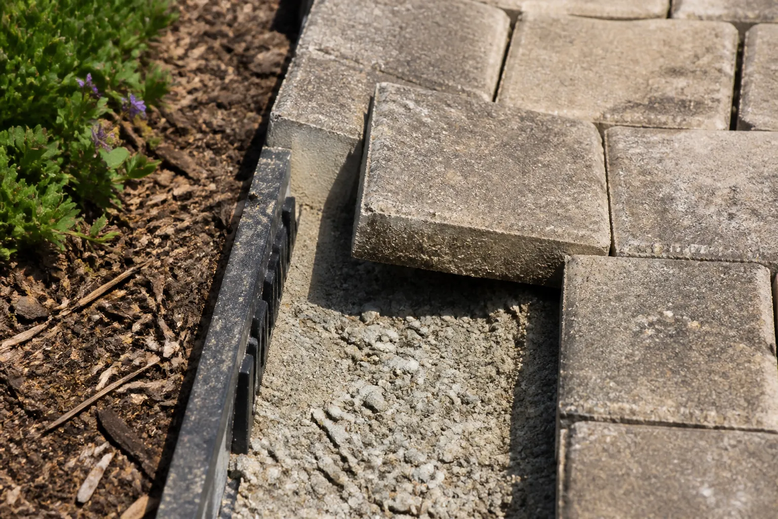A lifted paving block reveals the compacted base and drainage edge beneath a worn garden path.