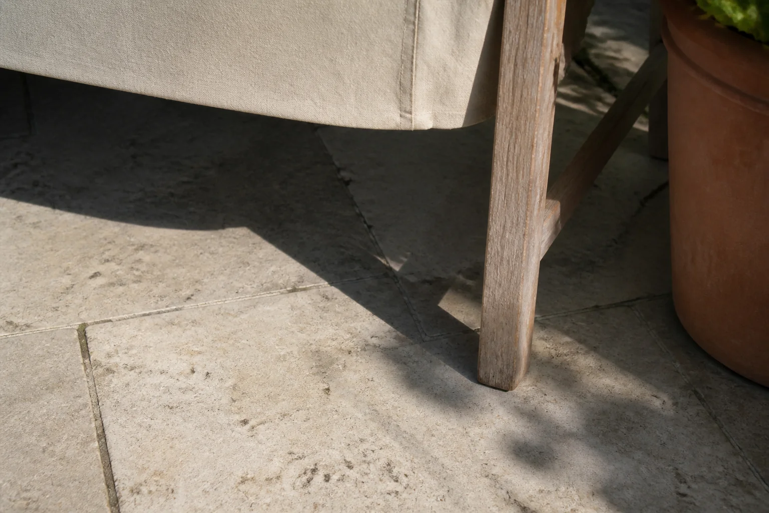 A worn wooden chair leg and canvas edge sit in useful shade beside a terracotta pot on pale paving.
