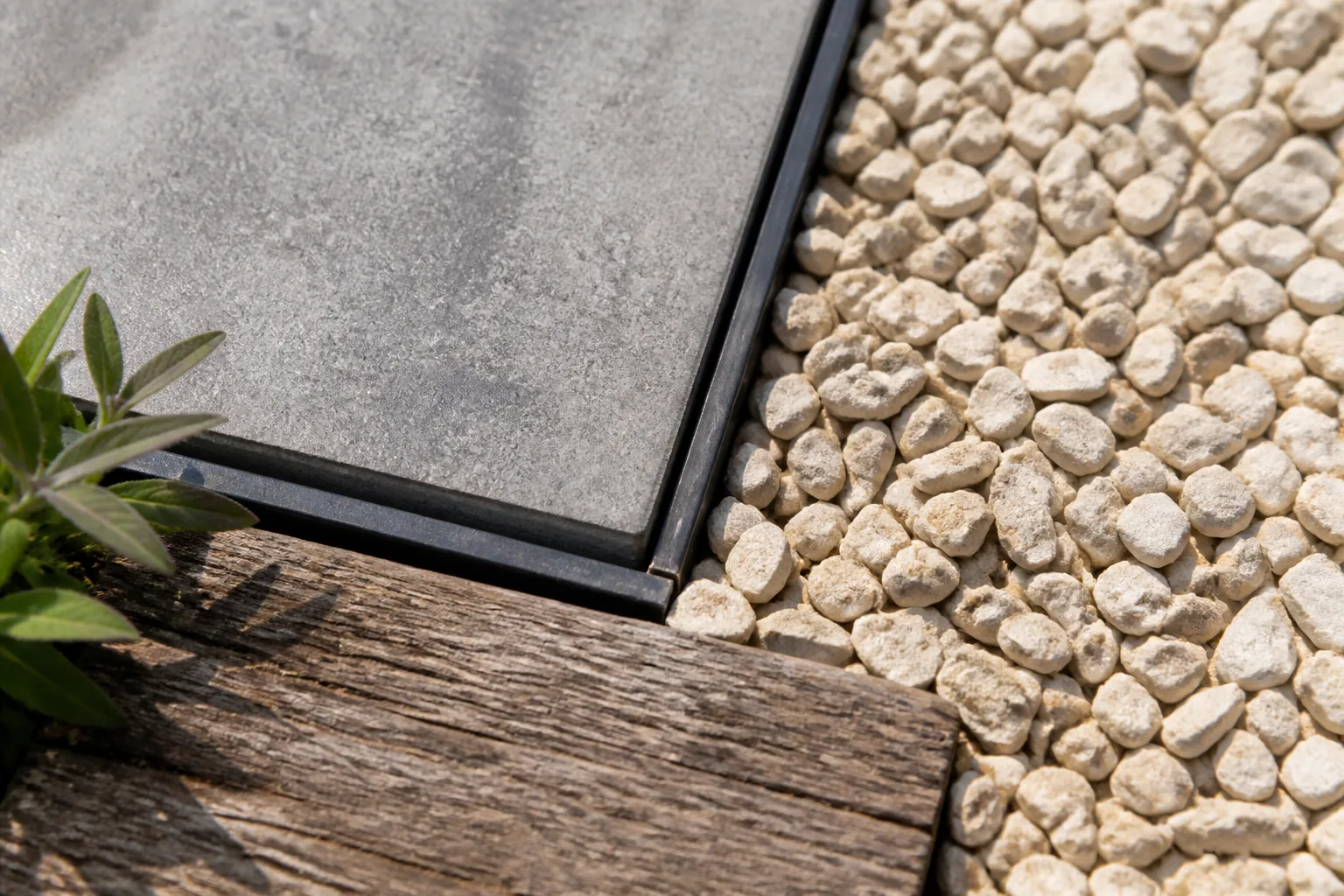 A grey concrete paver, pale gravel and weathered timber meet at a narrow dark steel edge.