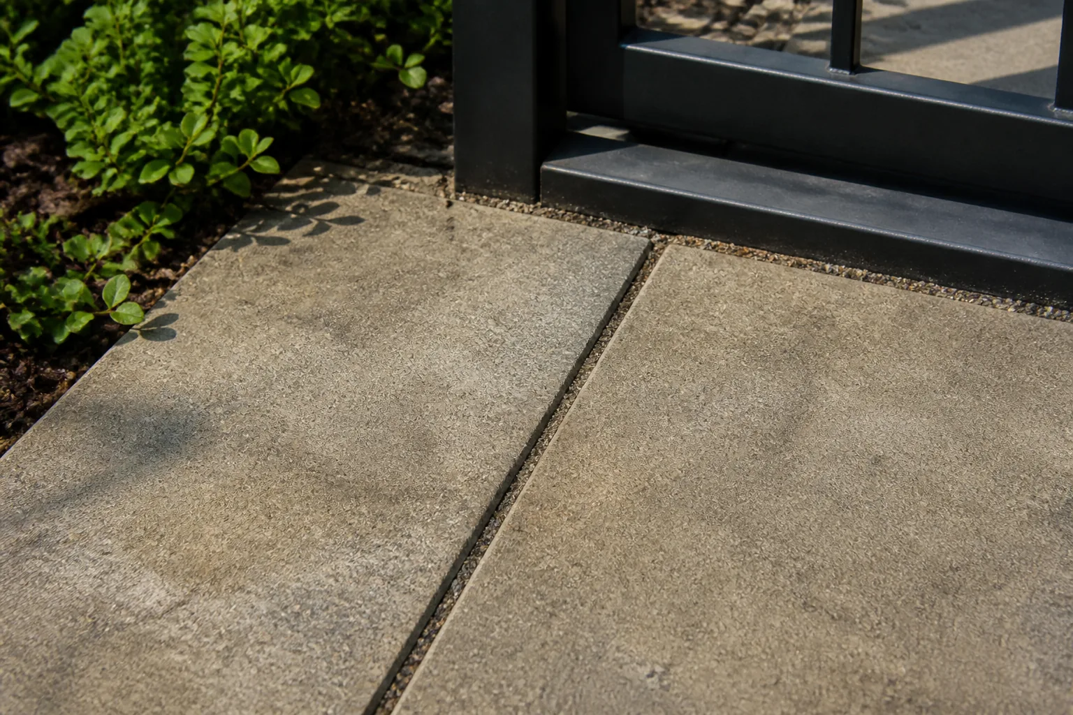 Slightly worn concrete pavers meet a dark metal gate threshold beside low green planting.