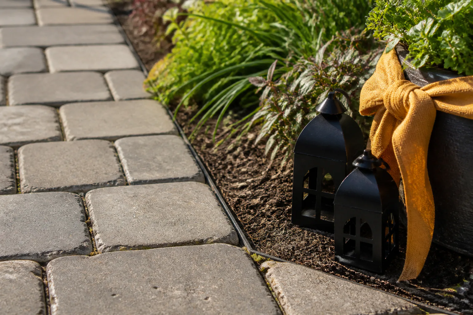 Two matte black lanterns and an ochre fabric accent sit in a planted edge, leaving the paved route clear.