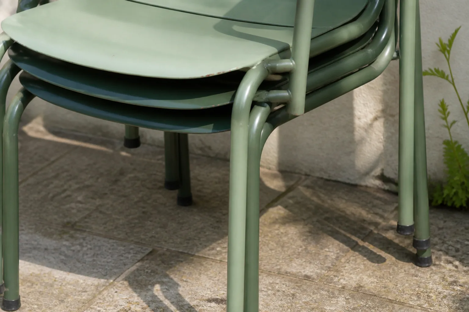 Three green metal patio chairs stack closely against a garden wall.