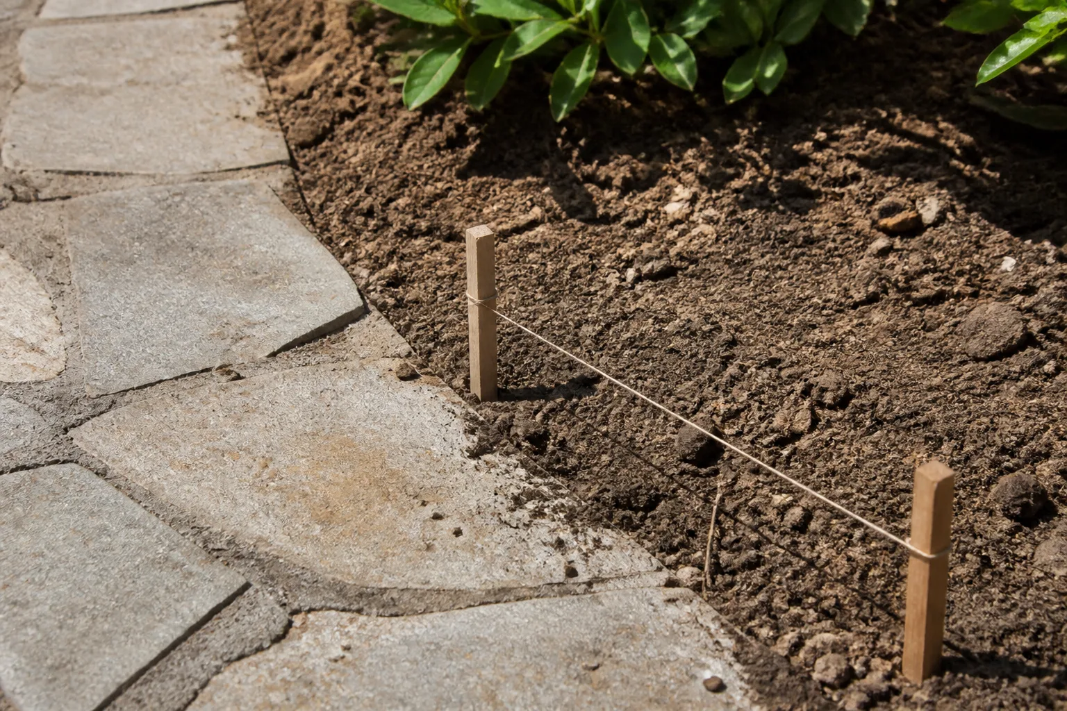 Houten paaltjes en een strak koord markeren een plantgrens naast een bestaand stenen pad voordat het werk begint.