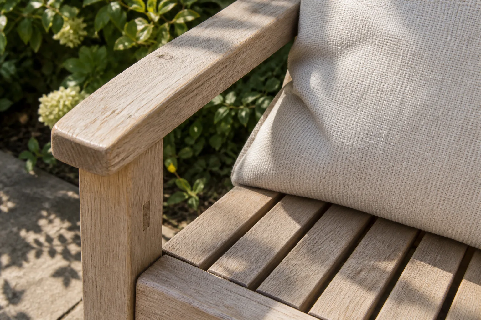 A pale weathered wooden chair arm beside a neutral cushion with visible woven texture.