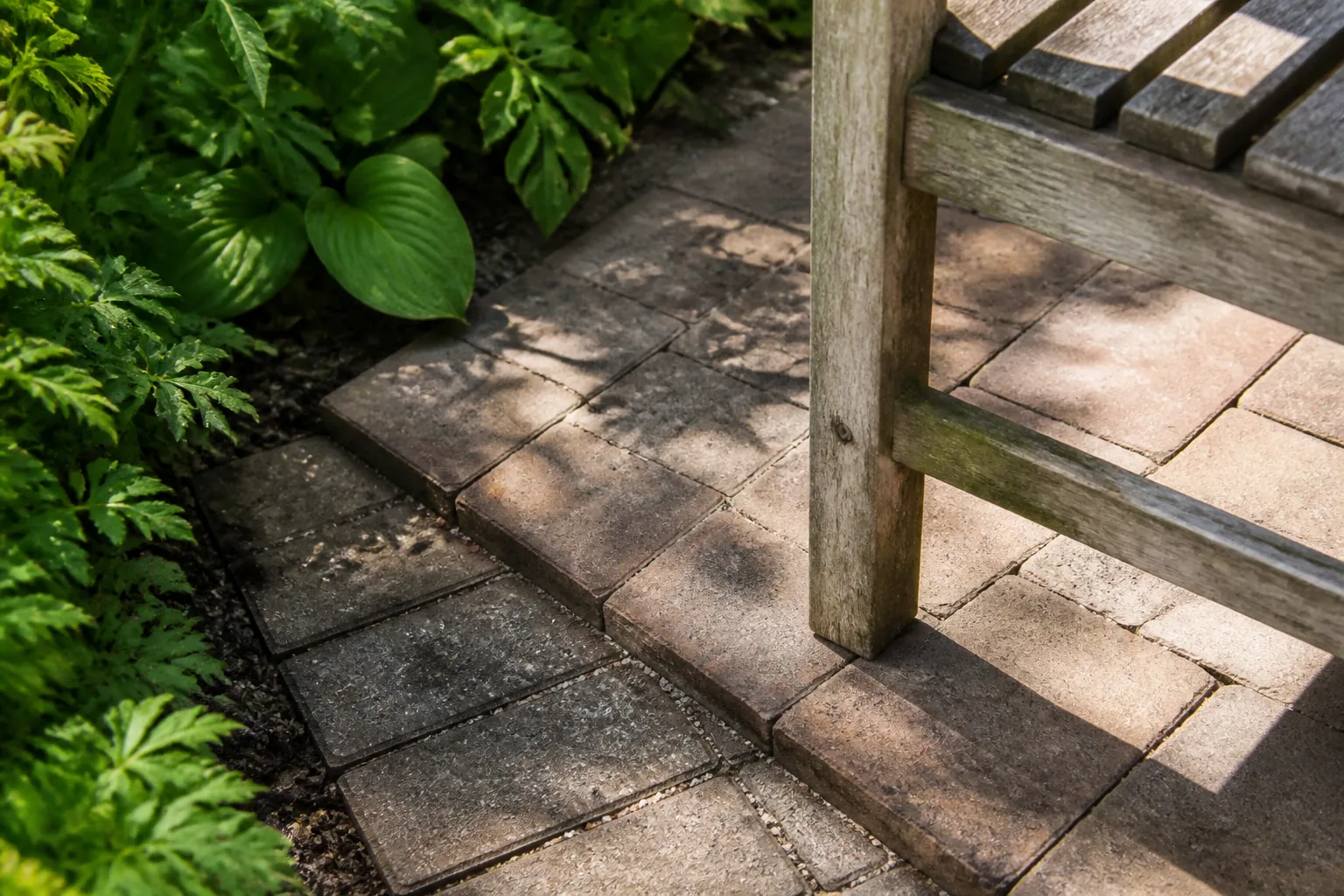 The leg of a weathered wooden chair stands where a narrow paver route widens beside a deep planted edge.