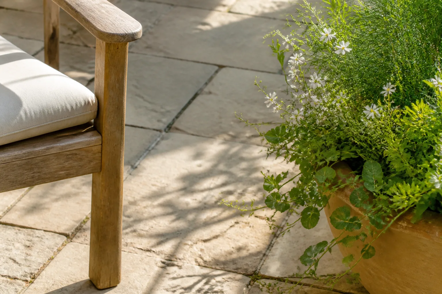 The arm and front leg of one comfortable wooden chair sit beside a generous planted container, with clear paving left between them.