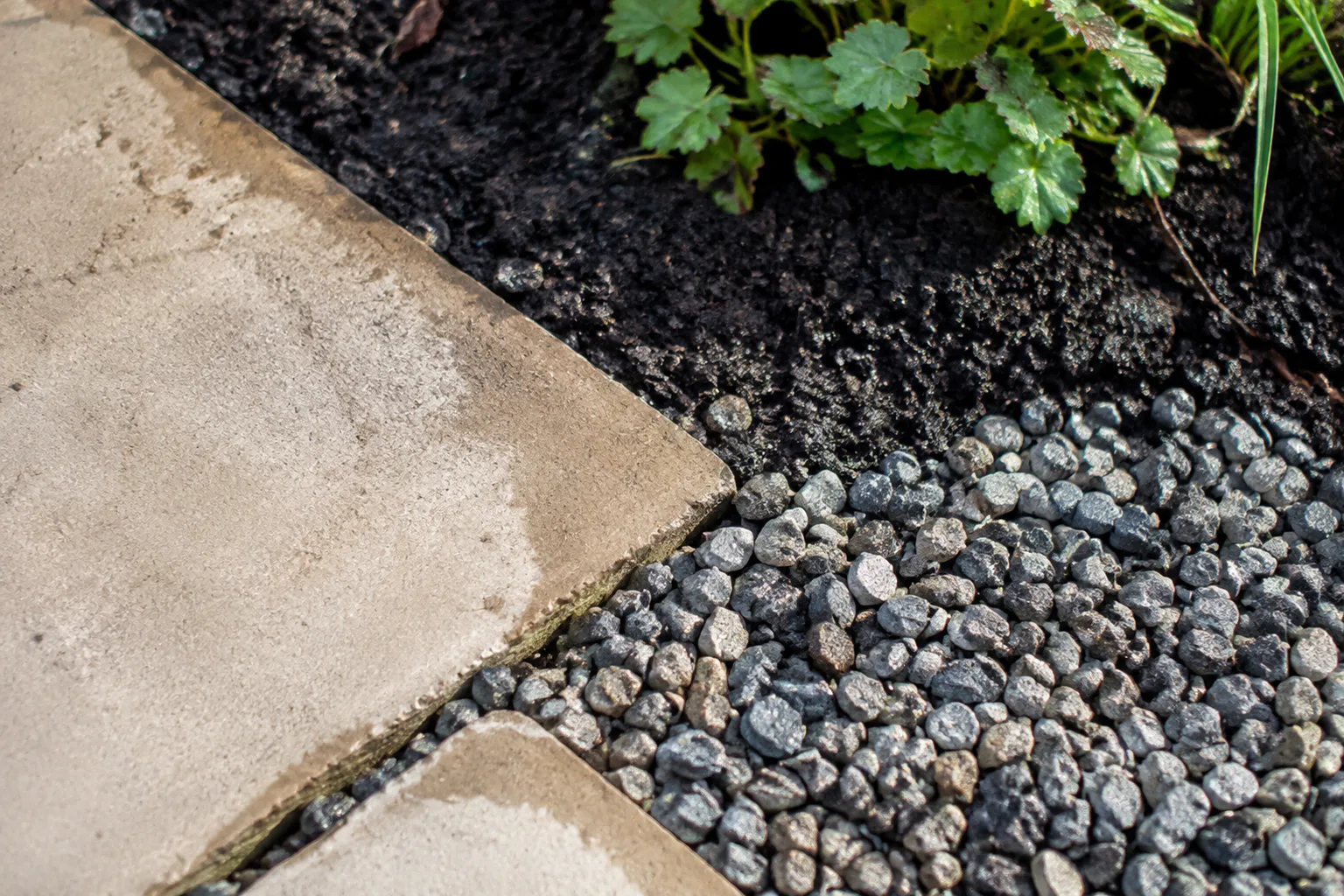 Een aansluiting van gebruikte bestrating, los grind en donkere aarde toont gewone vochtplekken zonder de rest van de tuin te laten zien.