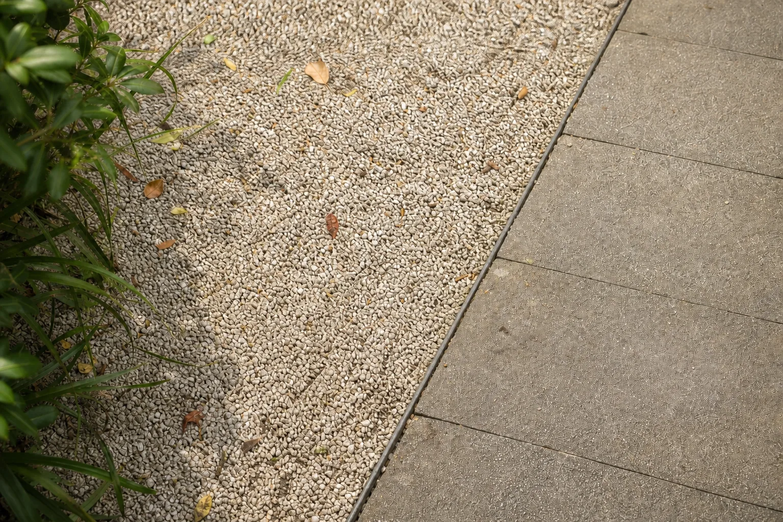 Pale gravel is held beside an existing paved path by a narrow dark edge, with a few fallen leaves showing the maintenance it will need.